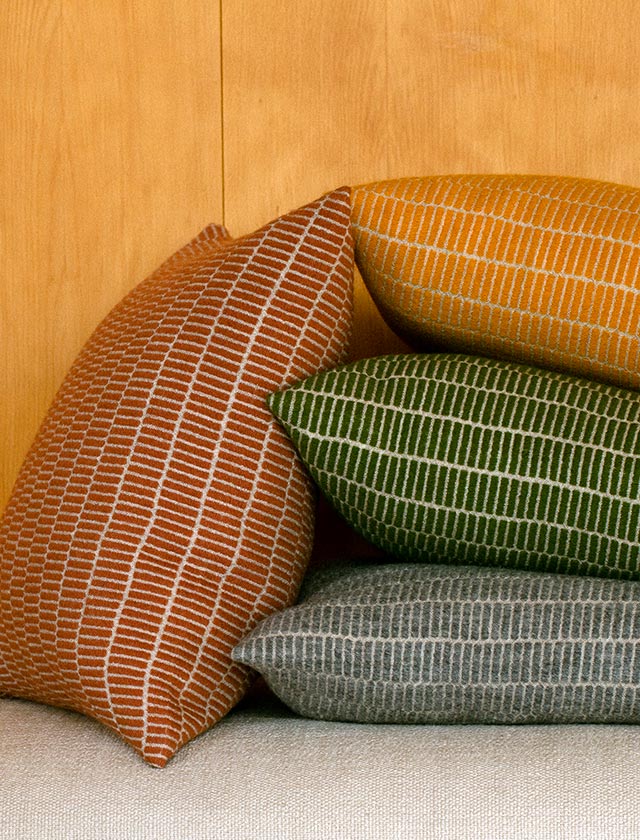 Vier handgewebte Kissenhüllen der RAITTI-Kollektion von Lapuan Kankurit aus reiner Wolle in den Farben Zimt, Gelb, Olive und Grau, übereinandergestapelt auf einem hellen Sofa vor einer Holzverkleidung – skandinavisches Wohnaccessoire mit natürlicher Textur.