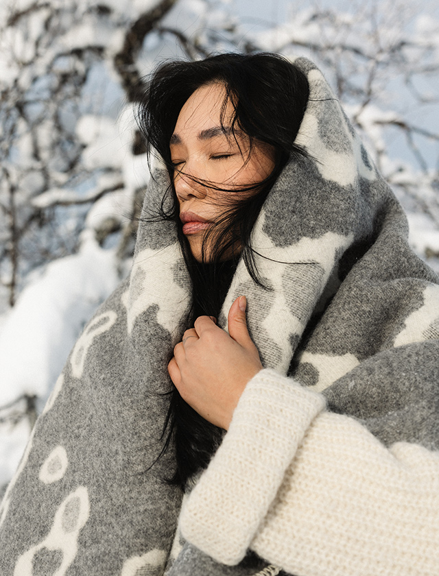 Frau im Schnee, eingehüllt in die weiß-graue Saimaannorppa Decke von Lapuan Kankurit.