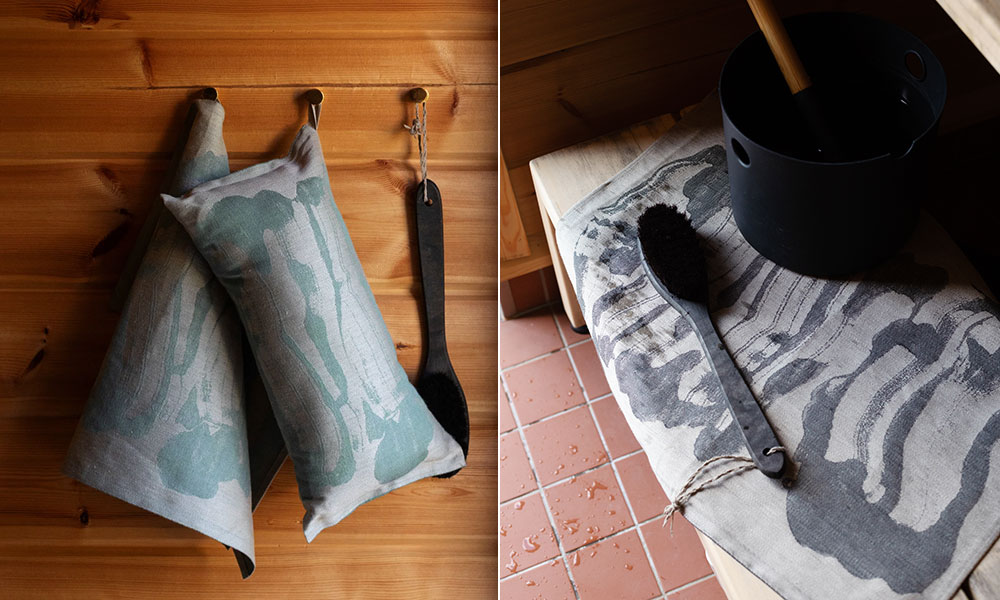 Savu Sauna-Serie der finnischen Weberei Lapuan Kankurit zu sehen: Zwei längliche Saunakissen aus Leinen hängen an Haken vor einer Holzsaunawand, daneben ein dunkler Massage- oder Reinigungsriemen. Rechts im Bild liegt eine Saunaauflage mit charakteristischem Rauchmuster auf einer Saunabank, ergänzt durch einen schwarzen Wassereimer mit Kelle und einer Bürste.