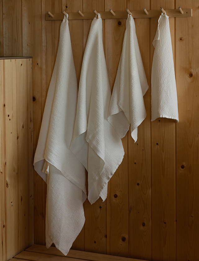 Lapuan Kankurit Terva Leinenhandtuch in Weiß, gefaltet als Stapel auf einer Holzbank vor hellem Fenster.