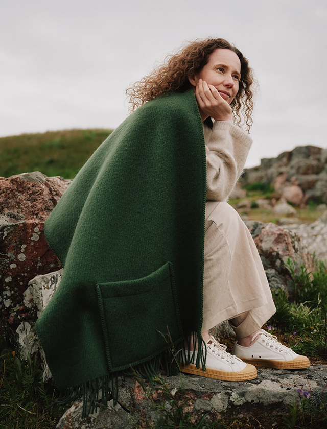 Eine Frau sitzt entspannt auf Felsen in der Natur und trägt das Lapuan Kankurit UNI Pocket Shawl in Dark Moss Green. Der wärmende Wollschal mit praktischen Taschen verbindet skandinavisches Design mit Funktionalität und ist ein stilvoller Begleiter für kühle Tage. Foto: Lina Jelanski.
