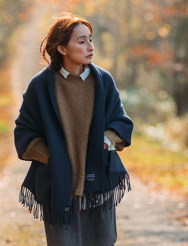 Eine Frau trägt den Lapuan Kankurit UNI Pocket Shawl in Midnight Blue an einem herbstlichen Waldweg. Der tiefblaue Schal mit Taschen und Fransen kombiniert nordisches Design mit praktischer Funktionalität. Gefertigt aus hochwertiger Wolle, eignet er sich ideal für Spaziergänge in der Natur wie auch für gemütliche Stunden zu Hause.