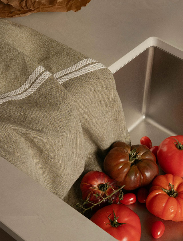 Küchentuch „Venla“ von Lapuan Kankurit – Klassisches Leinen in Oliv-Weiß Melange n einem schlichten Edelstahlspülbecken liegt ein Bündel frischer Tomaten, gebettet auf das edle Küchentuch „Venla“ von Lapuan Kankurit. Das Tuch in Oliv-Weiß Melange besticht durch seine natürliche Farbgebung und die feine Webstruktur aus reinem Leinen. Die charakteristische Fischgrät-Bordüre in Weiß verleiht dem Design eine klassische Note. „Venla“ ist nicht nur optisch ein Highlight, sondern überzeugt auch funktional – es ist saugfähig, langlebig und schnell trocknend. Ob beim Abtrocknen von Geschirr, zum Polieren von Gläsern oder als stilvolle Unterlage für frisches Gemüse: Dieses hochwertige Leinentuch vereint Ästhetik und Nachhaltigkeit auf höchstem Niveau und bringt skandinavisches Design in die moderne Küche.