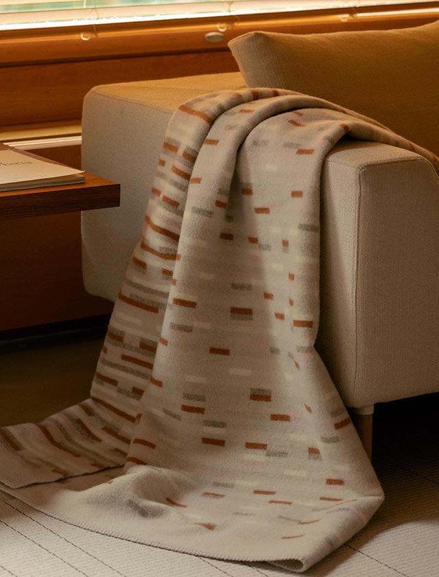 Gemütliche Szene in einem stilvoll eingerichteten Raum zu sehen. Im Vordergrund liegt eine hochwertige Wolldecke über die Armlehne eines beigen Sofas drapiert. Es handelt sich um die Decke „YLLÄTYS“ des finnischen Herstellers Lapuan Kankurit, bekannt für seine nachhaltige Produktion und traditionelle Webkunst im modernen skandinavischen Design.