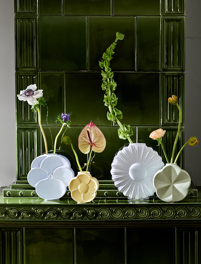Auf einem dunkelgrünen, glänzend gekachelten Sims stehen vier blumenförmige Porzellanvasen in Pastelltönen (Hellblau, Gelb, Weiß und Salbeigrün). In jeder Vase steckt ein einzelner, langer Blumenstiel mit zarten Blüten und Knospen in Weiß, Violett und Apricot sowie einem rosafarbenen Anthurienblatt. Die Szene wirkt minimalistisch und dekorativ – ein stilvolles Stillleben. Hersteller: Lyngby Porcelæn, Serie: Lyngby Flower Vasen.