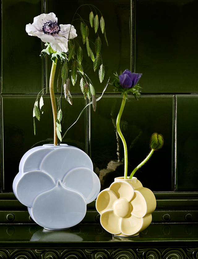 Zwei Lyngby Porcelæn Flower Vasen mit Stiefmütterchen und Butterblumen auf einem grünen Kaminsims vor dunkelgrüner Fliesenwand.