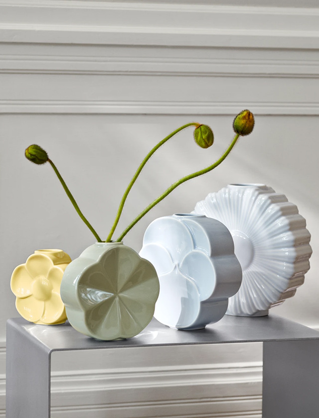 Flower Vasen-Serie von Lyngby Porcelæn: Vier blütenförmige Porzellanvasen in Pastellfarben (Gelb, Mintgrün, Hellblau und Weiß) stehen dicht arrangiert auf einem kleinen Tisch/Podest vor einer weißen Wand mit Zierleisten. In der grünen Vase stecken mehrere lange, gebogene Stiele mit Knospen – cleanes, skandinavisches Stillleben.