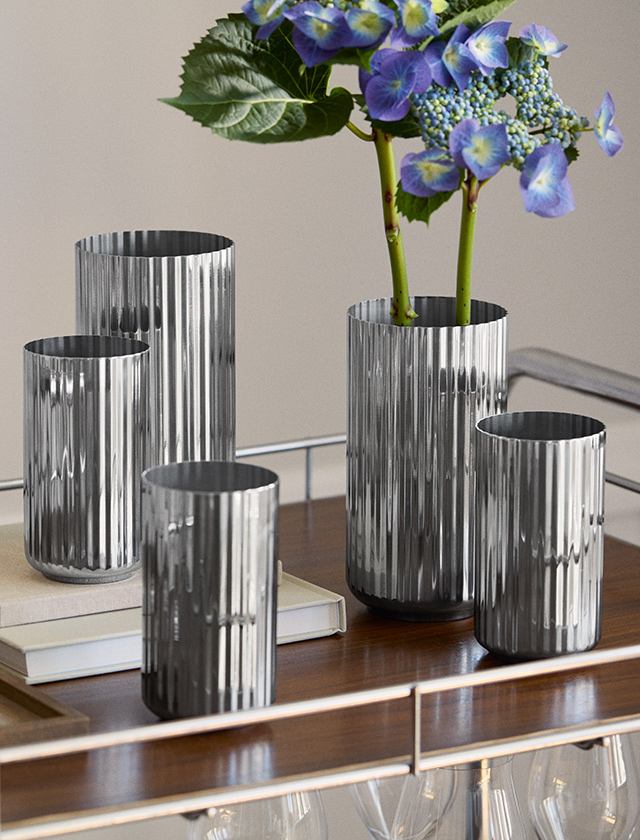 Lyngby Porcelæn Jubiläumsvase aus Edelstahl mit charakteristischer Rillenstruktur in verschiedenen Größen, dekoriert mit blauer Hortensie auf einem Design-Sideboard.