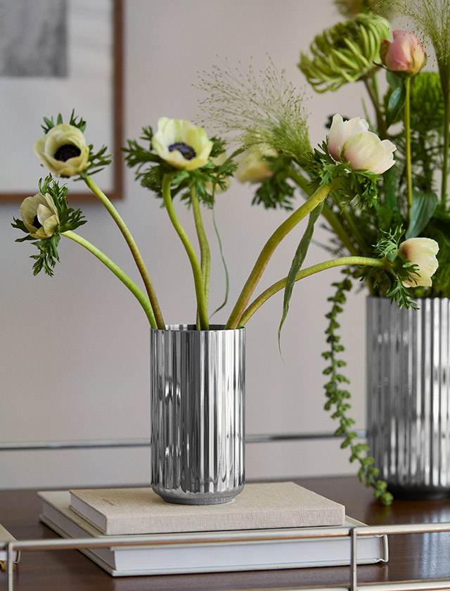 Lyngby Porcelæn Jubiläumsvase aus Edelstahl mit charakteristischer Rillenstruktur in zwei verschiedenen Größen, dekoriert mit filigranen Zweigen und Blüten auf einem Design-Sideboard.