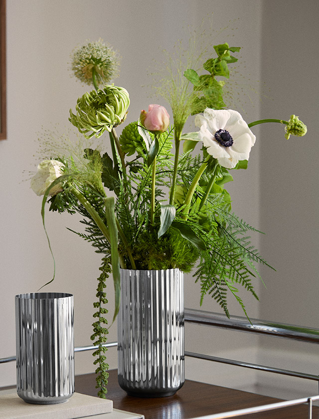 Lyngby Porcelæn Jubiläumsvase aus Edelstahl mit charakteristischer Rillenstruktur in zwei verschiedenen Größen, dekoriert mit üpigen Blüten und Zweigen auf einem Design-Sideboard.