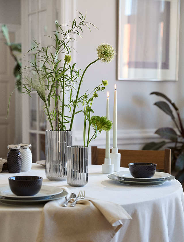 Zwei geriffelte Edelstahlvasen von Lyngby Porcelæn in 15,5 und 20,5 cm Höhe stehen mit filigranen Zweigen und Blüten auf einem elegant gedeckten Esstisch. Die silberfarbenen Vasen setzen einen klaren, skandinavischen Akzent zwischen Kerzen, Geschirr und dezenter Tischdekoration.