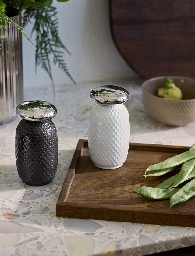 Lyngby Porcelæn Rhombe Salz- und Pfeffermühle aus Porzellan in Weiß und Schwarz mit glänzenden Metallaufsätzen, dekoriert auf einem Holztisch mit grünen Bohnen.
