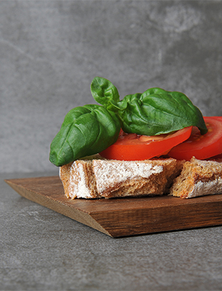 Close-up eines kleinen Brettes aus Eichenholz von Raumgestalt mit der praktischen abgeschrägten Kante. Auf dem Brett wird eine Scheibe Schwarzbrot mit Kruste serviert, belegt mit Tomatenscheiben und Basilikum.