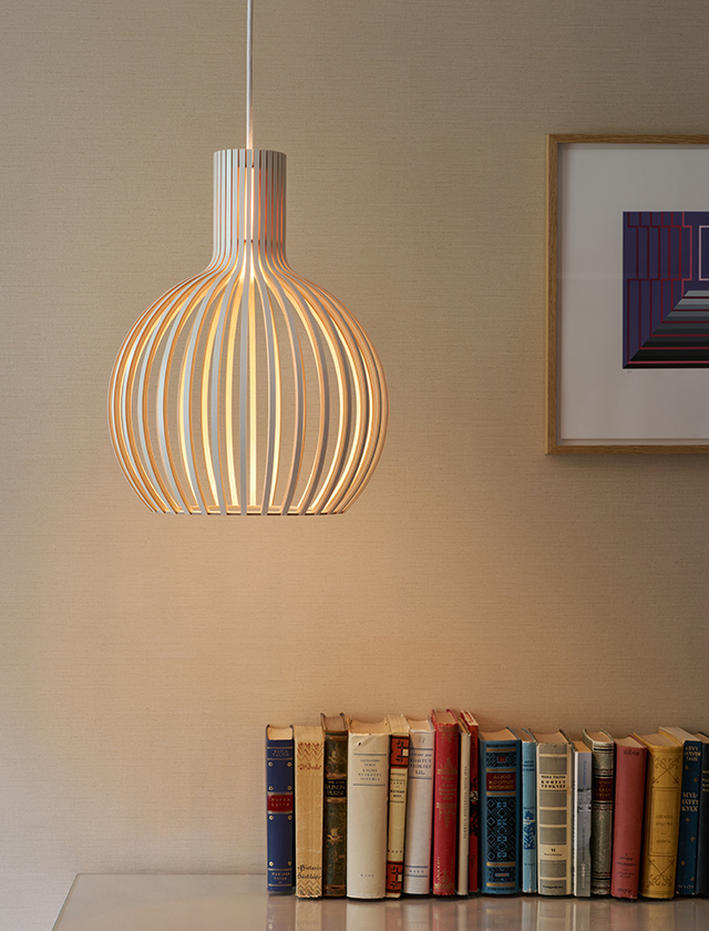 Secto Design Octo Tres 4242 Pendelleuchte in Weiß über einem Sideboard mit Büchern. Die Leuchte aus formgepressten Birkenlamellen erzeugt ein warmes, blendfreies Licht und zeigt die charakteristische offene Form der Octo Leuchtenfamilie.