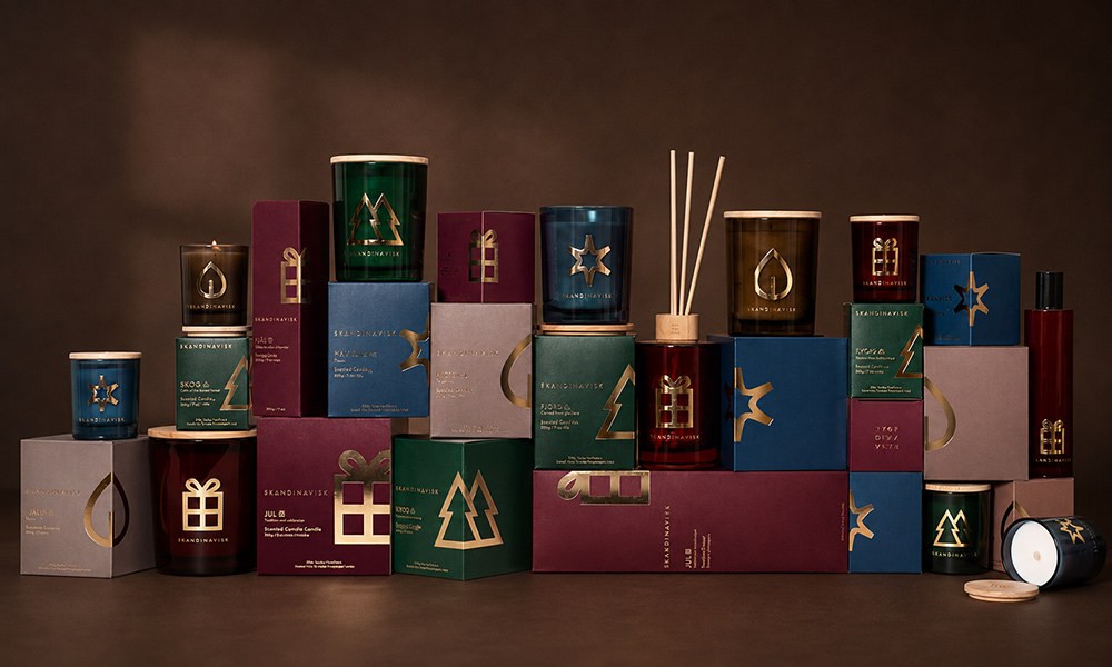Skandinavisk Duftkerzen, Diffuser und Geschenksets der Düfte JUL, SKOG, STJÄRNA, und HYGGE in festlicher Geschenkverpackung vor warmbraunem Hintergrund.