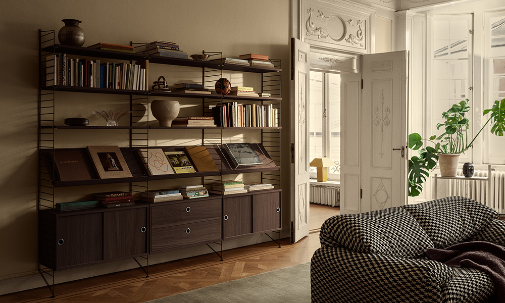 String Regal von String Furniture in dunkler Eiche (Dark Oak) als modulares Bücherregal im hellen Wohnzimmer, mit Büchern, Plattencovern, Pflanzen und Deko im minimalistischen skandinavischen Stil.