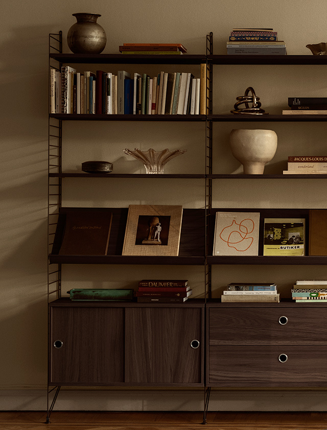 String Regalsystem von String Furniture mit dunkelbraunen Metallleitern und Regalböden aus dunkel gebeizter Eiche, bestückt mit Büchern, Keramik und Stauraummodulen mit Schiebetüren und Schubladen.