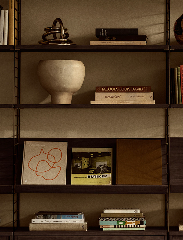 Das String Regal System aus dunkel gebeizter Eiche kombiniert Fachböden und Magazinablagen an filigranen Wandleitern. Die warme Inszenierung mit Büchern und ausgewählten Designobjekten betont den minimalistischen Wohnstil.