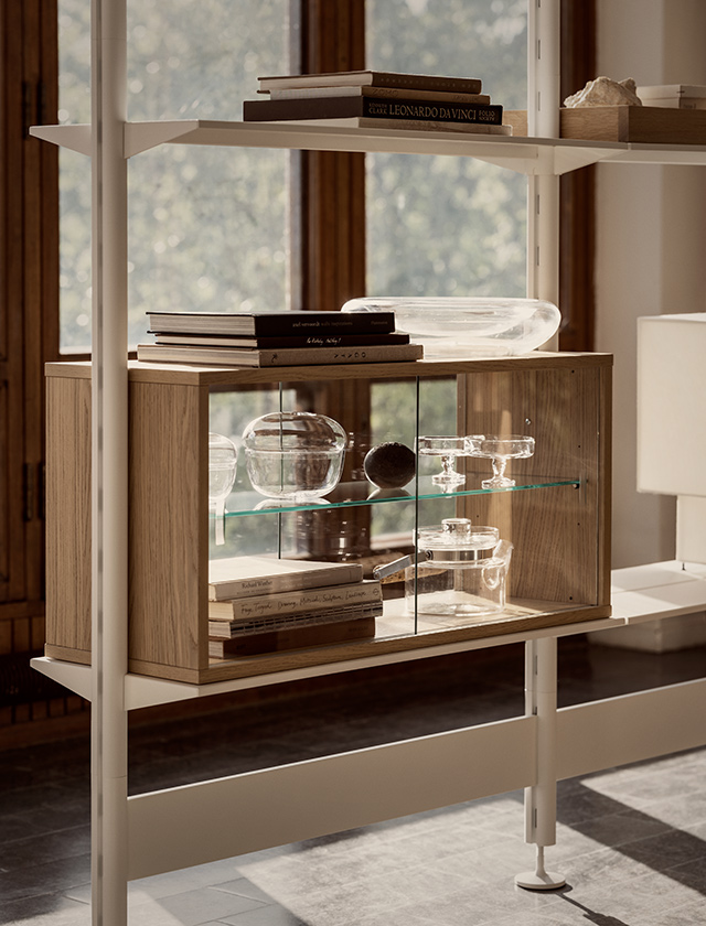 Das Pira Regal System von String Furniture in Eiche kombiniert offene Regalböden mit dem Vitrinenmodul CAB90 mit Glasboden und beidseitigen Glasschiebetüren (vorn und hinten). So lassen sich Glasobjekte, Bücher und Wohnaccessoires stilvoll präsentieren und von beiden Seiten bequem erreichen.