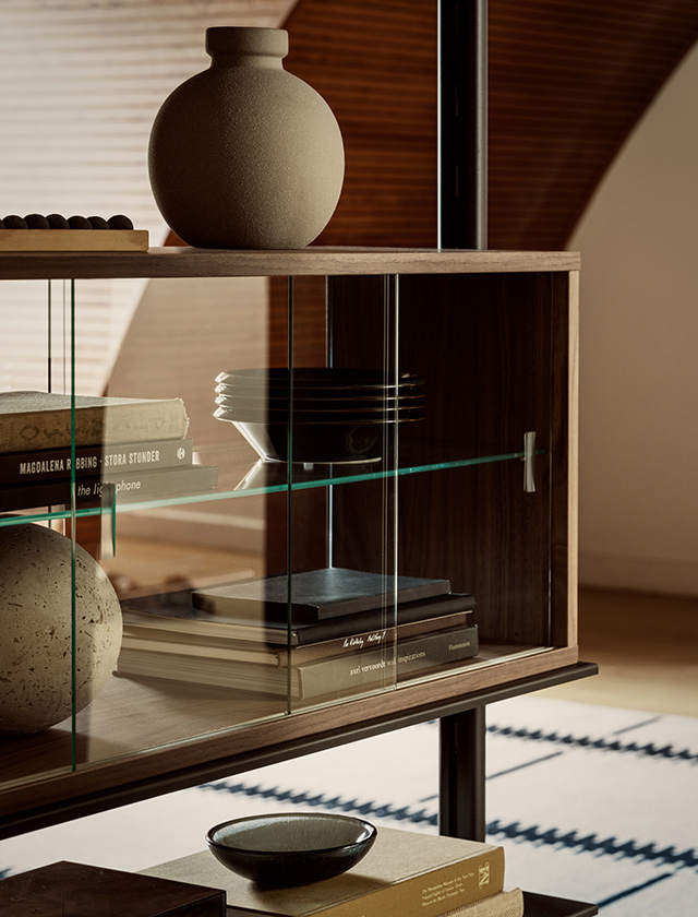 Nahaufnahme eines String Furniture PIRA Regalsystems: Vitrinenschrank Nussbaum mit Klarglas-Schiebetüren, innen Bücher und Geschirr, oben Vase und Schalen.