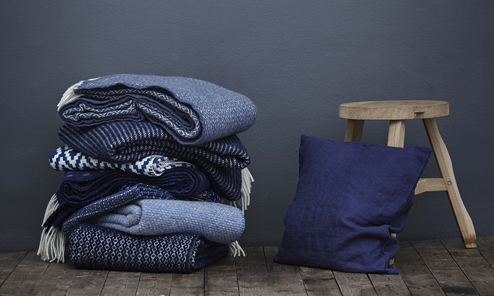 Das Foto zeigt einen Stapel Decken aus feiner Lammwolle und Baumwolle in blauen Farbtönen. Gefertigt werden die Wolldecken von Klippan Yllefabrik aus Schweden. Alle Decken weisen schöne skandinavische Muster auf. Neben dem Deckenstapel lehnt noch ein blaues Kissen an einem dreibeinigen Hocker.