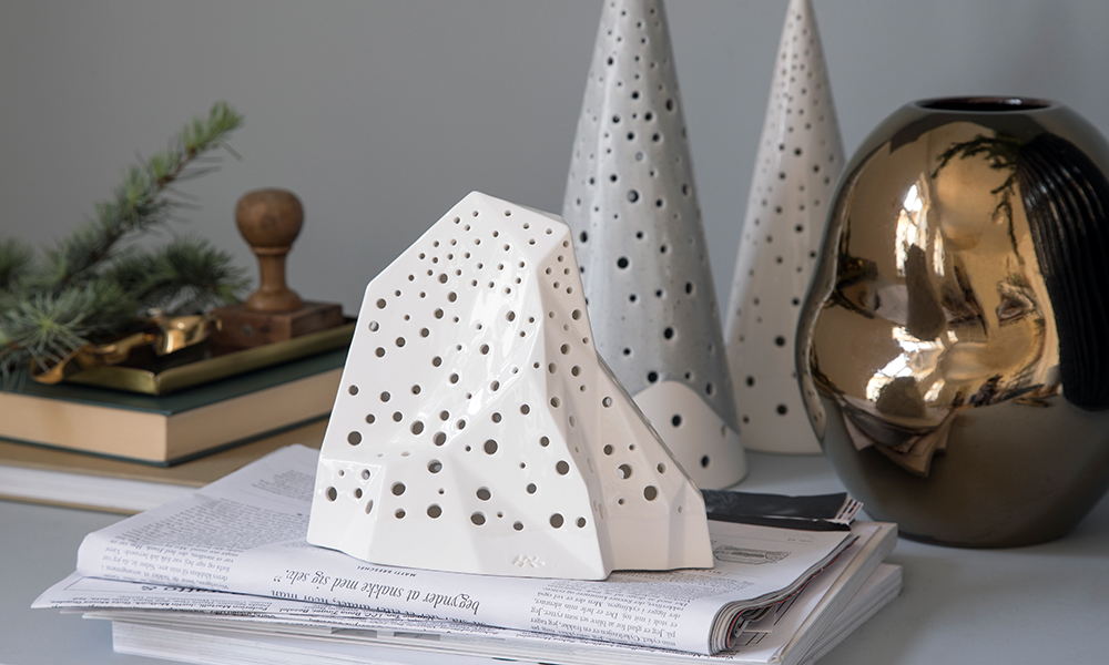 Weihnachtliches Arrangement aus zwei Nobili-Teelichtleuchtern, einem Nobili-Mountain-Teelichtleuchter und einer goldenen Fiora-Vase von Kähler Design. Der Nobili-Mountain steht leicht erhöht auf einem Stapel Zeitschriften und Journalen. Im Hintergrund ist ein Stapel Bücher mit Stempel und einem grünen Lärchenzweig zu sehen.